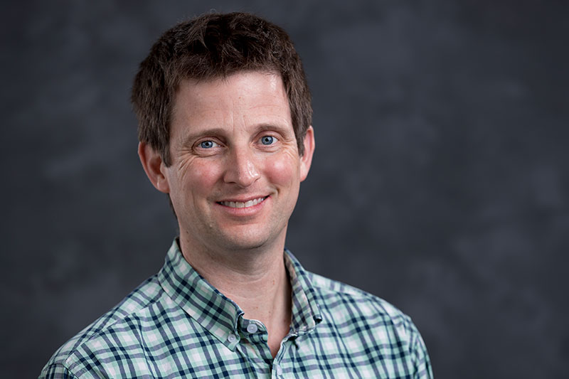 A person with short brown hair, wearing a green and white checkered shirt, smiles at the camera. The background is a dark, blurred grey.