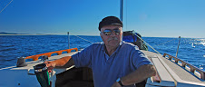 A man wearing a cap and sunglasses is sitting on a sailboat on a clear, sunny day. He is holding a cup and appears relaxed, with calm blue waters and a distant shoreline in the background.