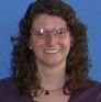 A woman with curly brown hair, wearing glasses and a purple top, smiles at the camera. The background is a solid blue color.