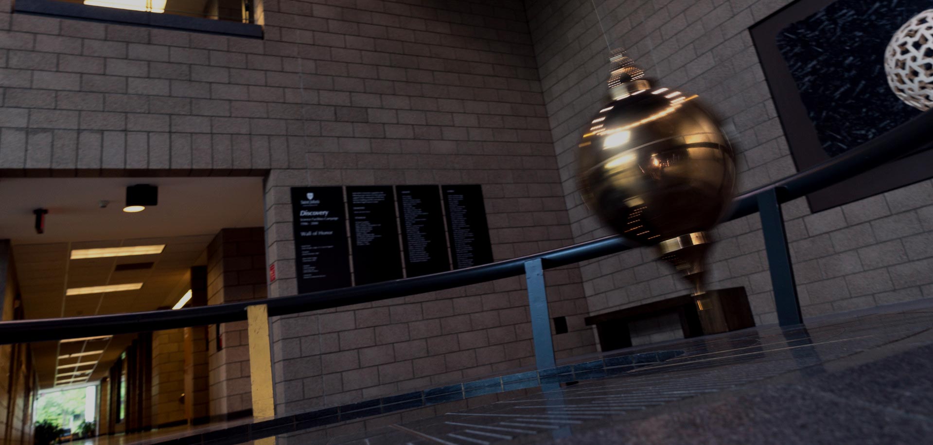 A large Foucault pendulum swings in a modern building with gray brick walls. Offices and informational displays are visible in the background. The pendulum's motion is captured mid-swing, showcasing its golden orb.