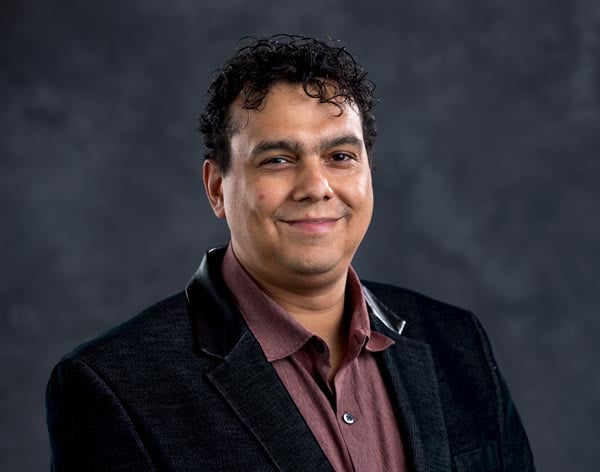 A man with curly dark hair is standing against a dark background, wearing a dark jacket with a shiny black collar detail and a maroon button-up shirt. He is smiling slightly while looking at the camera.