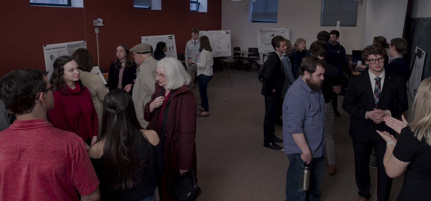 A group of people is gathered in a room, engaged in discussions and presentations. Various posters are displayed on stands behind them. The attendees are spread out in small groups, some standing and talking, some closely examining the posters.
