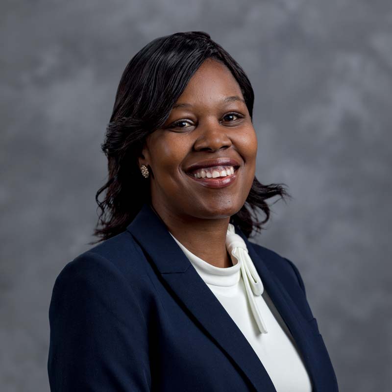 A person with medium-dark skin tone, shoulder-length black hair, and a bright smile is wearing a dark blue blazer over a white blouse. They are posing against a grey, slightly textured backdrop.