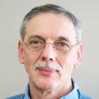 A middle-aged person with short gray hair, glasses, and a mustache, wearing a light blue collared shirt, looks directly at the camera against a plain background.