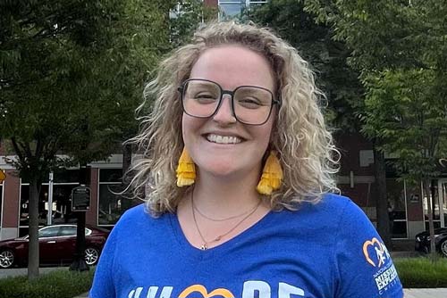 A smiling person with curly blonde hair and glasses is wearing yellow earrings and a blue shirt that says "Hope" with a heart logo. Trees and buildings with a parked red car are visible in the background.