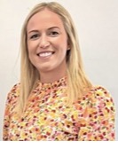 A woman with long, blonde hair smiling at the camera. She is wearing a floral blouse with a high, ruffled neckline, and is standing against a plain, light-colored background.