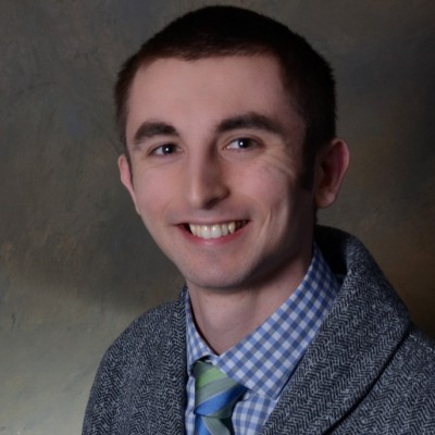 A young man with short brown hair is smiling at the camera. He is wearing a gray blazer over a blue and white checked shirt paired with a green and blue striped tie. The background is a plain, textured wall.