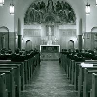 A monochrome image shows an interior view of a church. People are seated in rows of pews facing the altar. The ornate altar features a statue and religious artwork. The church has arched ceilings and lantern-style chandeliers hanging above.