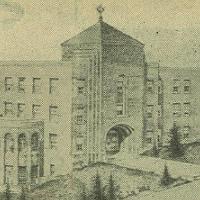 A sepia-toned image of a large, multi-story institutional building with a central tower featuring an arched entrance. The structure has columns and several small, evenly spaced windows. The surrounding area shows a few scattered trees and minimal landscaping.