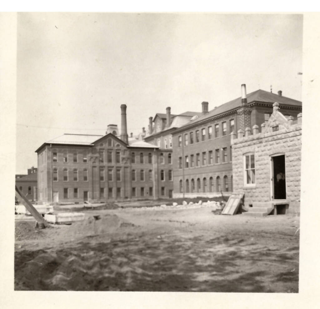 A black-and-white photograph of a large institutional building with multiple sections and several chimneys. The surrounding area appears to be a construction site with dirt and some building materials scattered around. A stone structure is visible in the foreground.