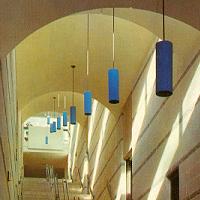 A hallway with a high arched ceiling featuring several cylindrical blue hanging lights. The walls and ceiling are light-colored, and the corridor is well-lit with natural light streaming in from windows off-frame. Stairs lead upwards at the end of the hallway.