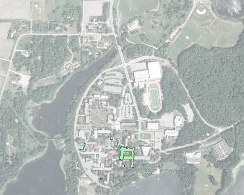 Aerial view of a large campus surrounded by trees and water. Multiple buildings and a sports field with a track are visible. Pathways and roads connect different areas within the campus. A prominent green-roofed building is located centrally.