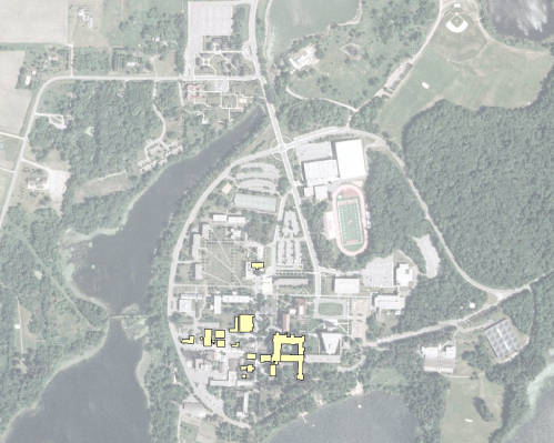 Aerial view of a campus surrounded by trees and a lake. The image shows various buildings, pathways, and a stadium with a track. The area is lush with greenery and appears to be a large educational institution.