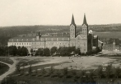 A historic, large brick building with two tall spires, surrounded by trees and a wide grassy area with people walking, set against a backdrop of forested hills under a gray sky.