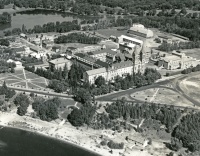 Aerial view of an expansive campus featuring several large buildings surrounded by trees, pathways, and open spaces. A body of water can be seen in the background alongside a prominent road and beach in the foreground.