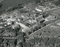 Aerial black-and-white photo of a large complex of interconnected buildings with a U-shaped courtyard, surrounded by trees and roads. A river is visible at the top. The architecture appears traditional, suggesting an institutional setting.