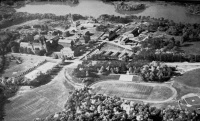 Aerial black and white photograph of a large campus surrounded by trees and a river. Buildings are scattered throughout, with a prominent central structure. Open fields and a sports track are visible in the foreground.