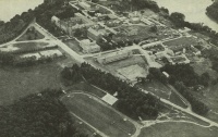 Aerial view of a campus with multiple buildings, tree-lined streets, and open fields. A running track and sports field are visible in the foreground. The area is surrounded by lush greenery and a body of water is seen in the background.