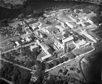 Black and white aerial view of a large, historic industrial complex with multiple buildings surrounded by trees and a river in the background. The scene shows roads, greenery, and industrial structures.