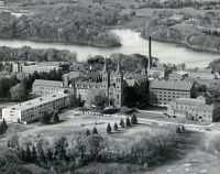 Aerial view of a large historic building complex with multiple sections, surrounded by trees and open fields. A river flows in the background. The architecture includes towers and a smokestack, giving a sense of an institutional or educational facility.