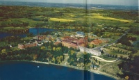 Aerial view of a large historic campus with red brick buildings surrounded by lush greenery and trees, bordered by a lake. Roads and fields stretch into the horizon under a clear blue sky.