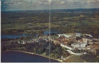 Aerial view of a large building complex surrounded by lush green fields and dense forests, with a river flowing nearby. A serene lake is visible in the foreground, under a partly cloudy sky.