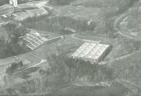 Aerial view of a landscape featuring a large building with a segmented roof surrounded by trees. Nearby, tiered structures resembling bleachers or steps are visible. A river flows through the lower part of the image.