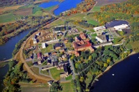 Aerial view of a large, complex facility surrounded by a bend in a river. Numerous red and white buildings, green lawns, and tree-lined paths are visible. The surrounding area features fields and patches of autumn-colored trees.