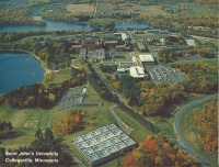Aerial view of Saint John's University campus in Collegeville, Minnesota, surrounded by autumn foliage. Several large buildings, roads, and a lake are visible in the landscape.
