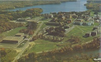 Aerial view of a college campus surrounded by trees and water. The campus features several buildings, open green spaces, and parking areas. There is a river in the background and the area is lush and green.