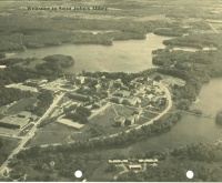 Aerial view of a large building complex surrounded by trees and several lakes. The landscape features winding roads and various structures, indicating a settled area within an expansive natural environment. The image has a vintage appearance.
