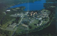 Aerial view of a large campus surrounded by lush greenery and several buildings, set near a blue lake. The landscape includes winding roads and open fields.
