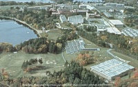 Aerial view of a large campus with multiple buildings surrounded by trees and a lake. The landscape shows a mix of greenery and autumn-colored foliage. Pathways connect the structures, and open fields are visible throughout the area.