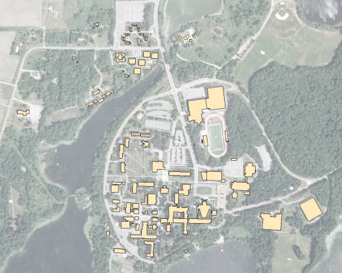 Satellite view of a large campus with various buildings marked in yellow, surrounded by trees and greenery. A river or lake is visible on the left, and there is a running track to the right. Roads and parking areas are visible throughout the area.