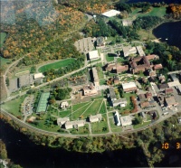 Aerial view of a campus surrounded by trees and bordered by water. Buildings are spread out with green spaces and pathways in between. The landscape is tinged with autumn colors, with a river visible on two sides.