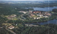 Aerial view of a large campus surrounded by lush greenery and several bodies of water. The campus consists of multiple buildings and roads, set amidst a forested landscape with lakes visible in the background.