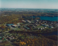Aerial view of a campus surrounded by trees and water. The area features several buildings, fields, and pathways, with a lake visible to the right. The landscape is lush and green, indicating a natural and serene environment.