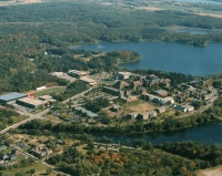Aerial view of a landscape featuring a large campus with numerous buildings surrounded by lush greenery and bodies of water. Roads and pathways connect the buildings, and a river flows in the foreground. Forested areas extend into the distance.