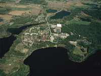 Aerial view of a sprawling complex surrounded by dense forests and multiple bodies of water. Roads intertwine through the landscape, with open fields and vegetation visible throughout. A large white building stands out in the center.