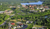 Aerial view of a lush, green landscape with a cluster of buildings, surrounded by dense forests and a river in the background. The image shows a mix of modern and older architecture, roads, and open spaces.