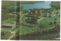 Aerial view of an expansive university campus with several buildings surrounded by green lawns, trees, and a body of water in the background. Paths and roads connect different structures, creating a structured layout.