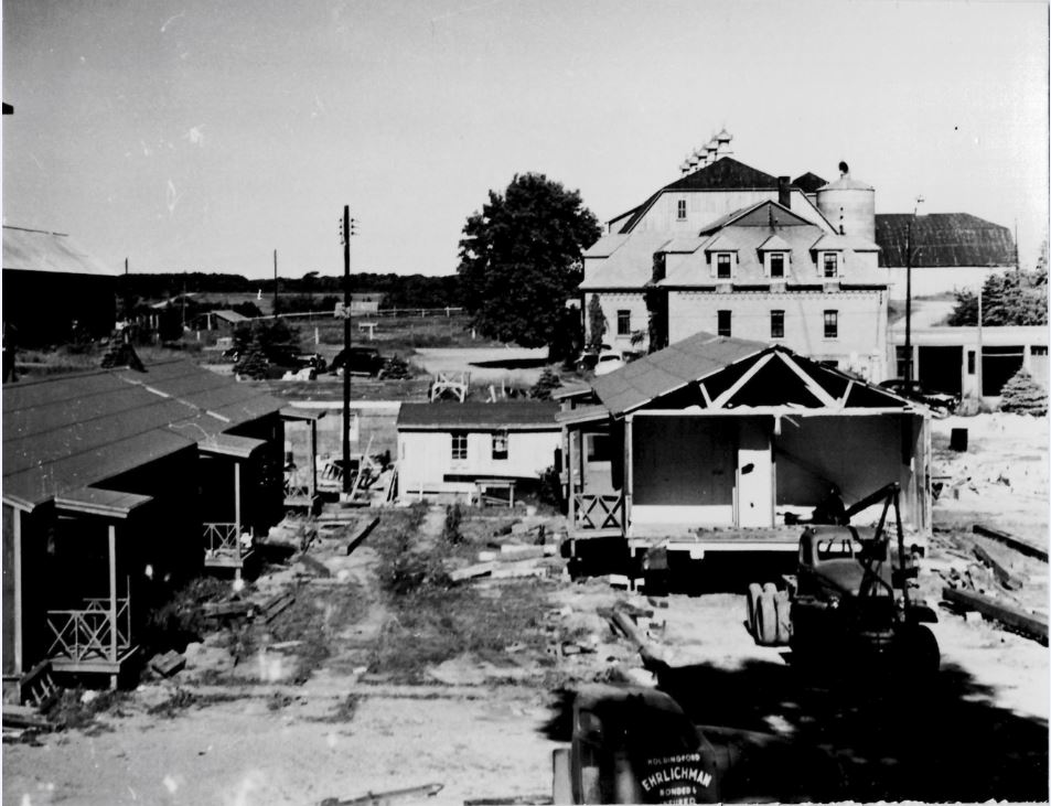 A black and white photo of a construction site with partially built structures. A partially assembled building is in the center. In the background, an older multi-story house and other structures are visible. Scattered building materials are around.