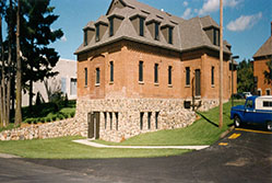 A two-story brick building with a stone foundation and triangular roof windows. It's surrounded by green grass and trees. A blue truck is parked on a paved road nearby under a clear blue sky.