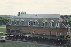 A large brick house is being transported on multiple wheels across a grassy area. A truck is parked nearby. The house has a steep roof with dormer windows and a brick exterior. Trees are visible in the background.