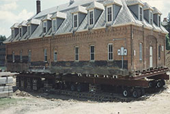 A large brick house is being relocated using a platform with wheels. The structure sits elevated on a series of beams and supports. The sky is clear, and there is a building partially visible in the background.