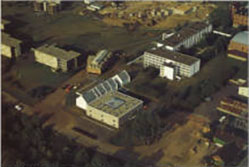 Aerial view of a campus with several rectangular buildings surrounded by greenery. A distinct building with a glass roof is central in the foreground, while other buildings are scattered around, and some construction appears in the background.