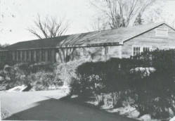 A black and white photo of a long, single-story building with a sloped roof, surrounded by bushes and trees. The foreground shows a road, and the leafless trees cast shadows on the structure.