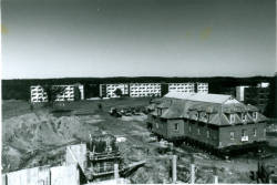 A black-and-white photo of a construction site with a large building under construction in the foreground. Several completed apartment blocks are visible in the background, situated in an open area with sparse vegetation.