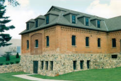A historic building with a stone foundation and red brick upper walls, featuring multiple dormer windows on its pitched roof. The structure is surrounded by green grass and trees under a cloudy sky.