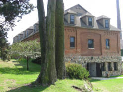 A large, two-story brick building with multiple dormer windows sits surrounded by grass and trees. In the foreground, two tall trees stand closely together. The building has a stone base and a sloped roof.